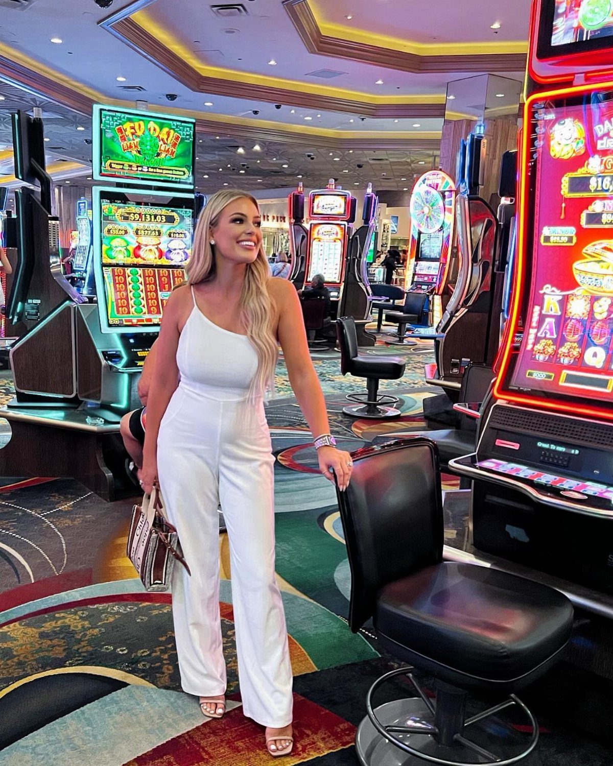 A blonde woman in a white jumpsuit posing inside a brightly lit casino.
