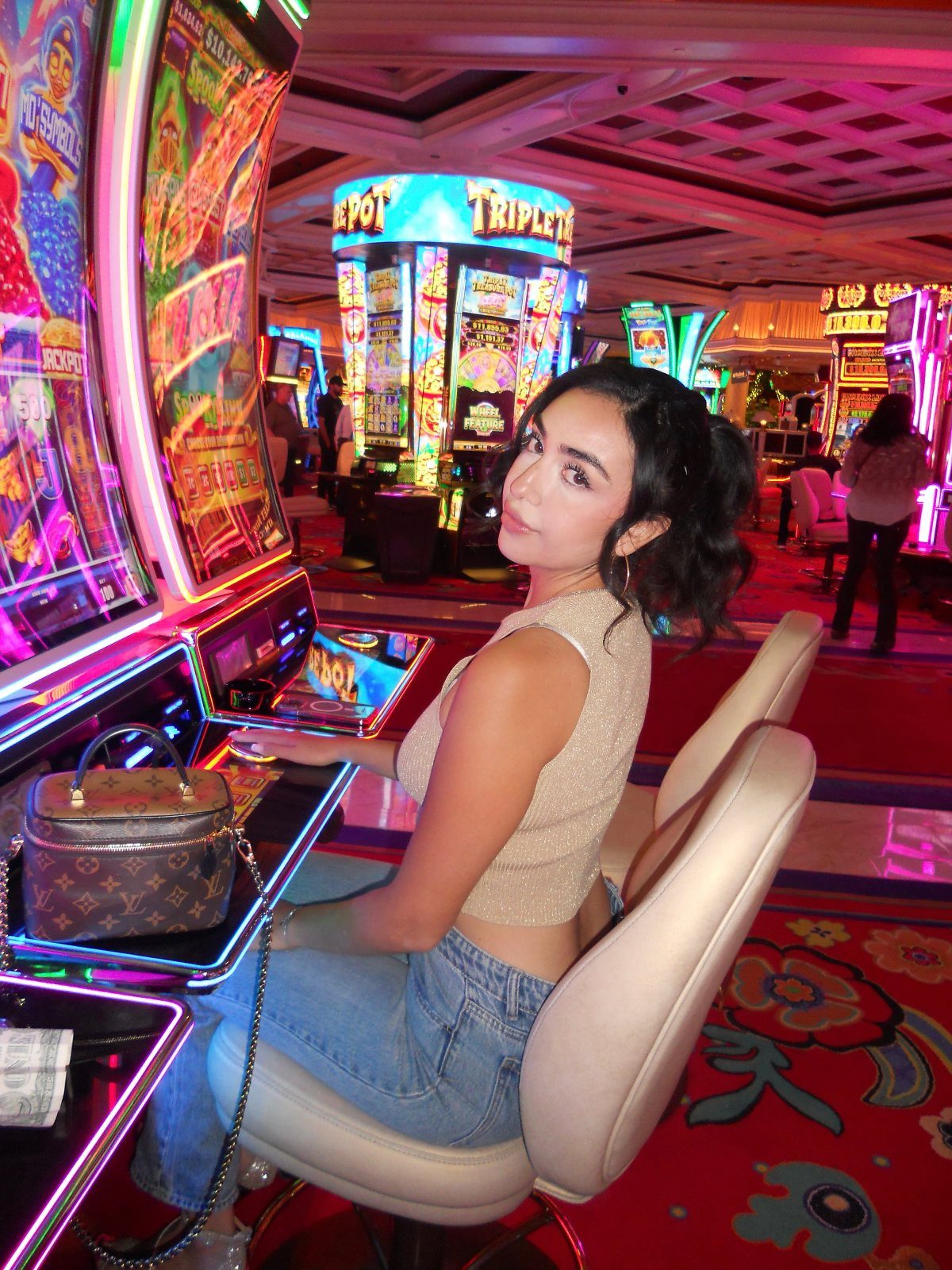 Young woman in a sparkly top sitting at a slot machine in a colorful casino.