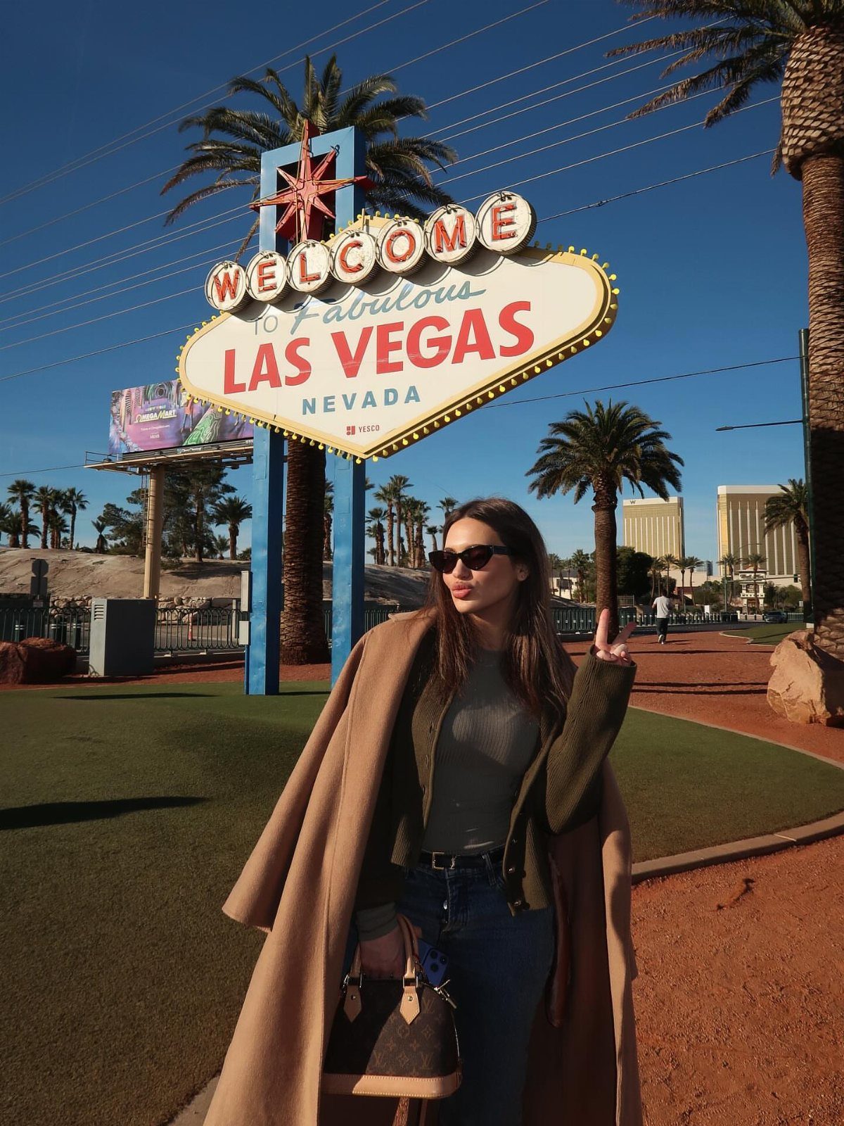 Woman in a tan coat and sunglasses posing in front of the Las Vegas sign.