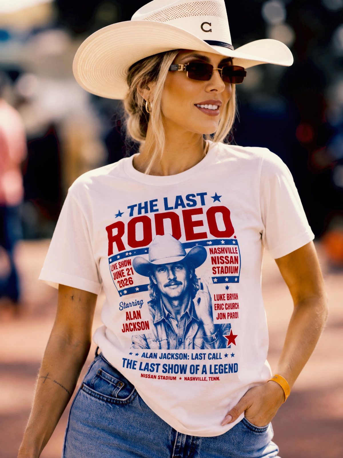 Woman in a white graphic t-shirt, blue jeans, and a white cowboy hat smiling outdoors.