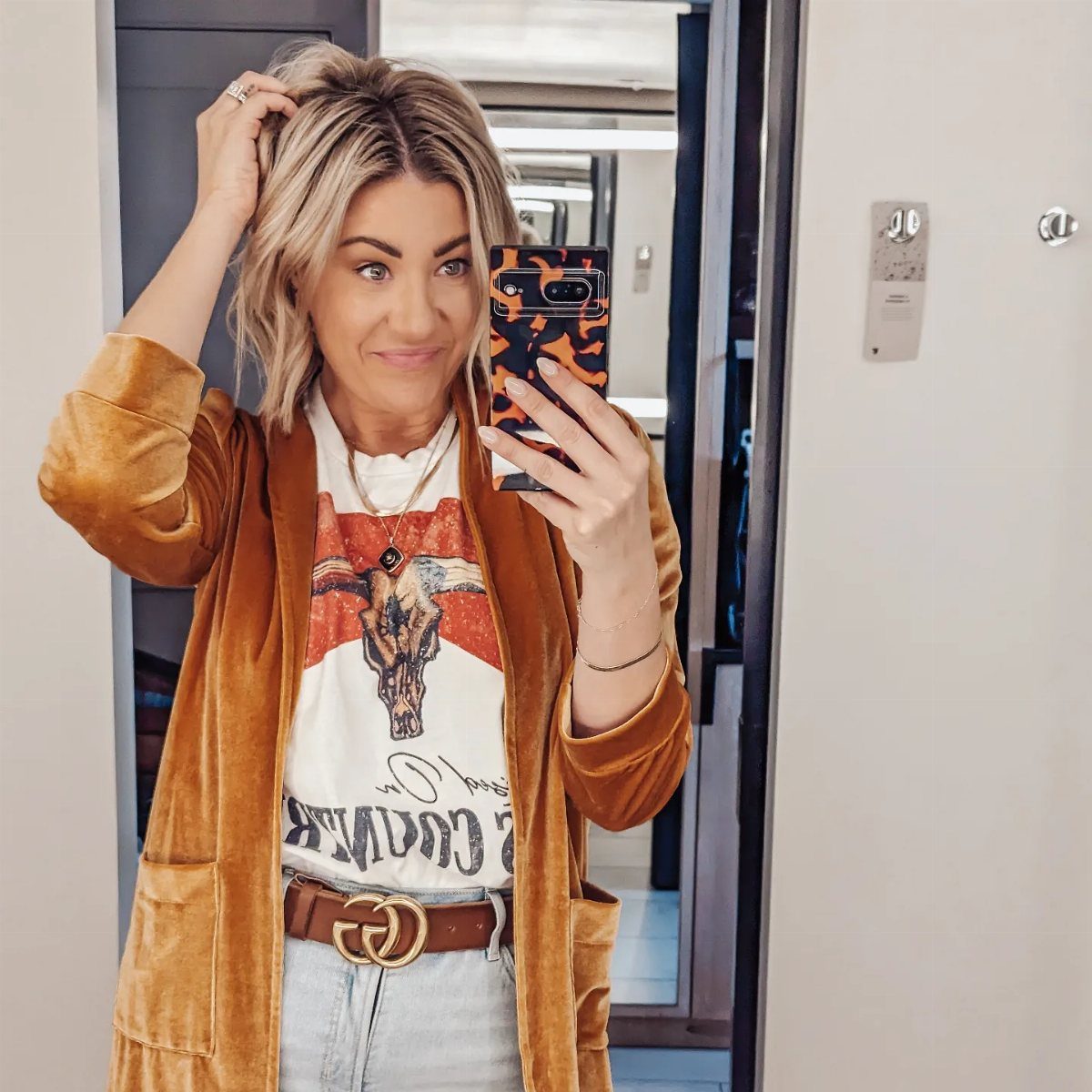 Blonde woman posing in a mirror wearing a graphic tee, velvet cardigan, and denim jeans.