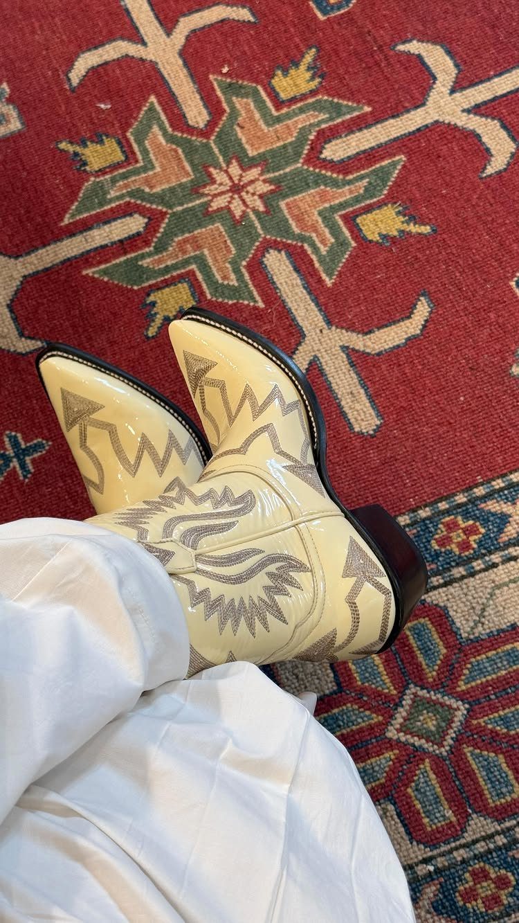 Cream embroidered cowboy boots resting on a red patterned rug next to white clothing.