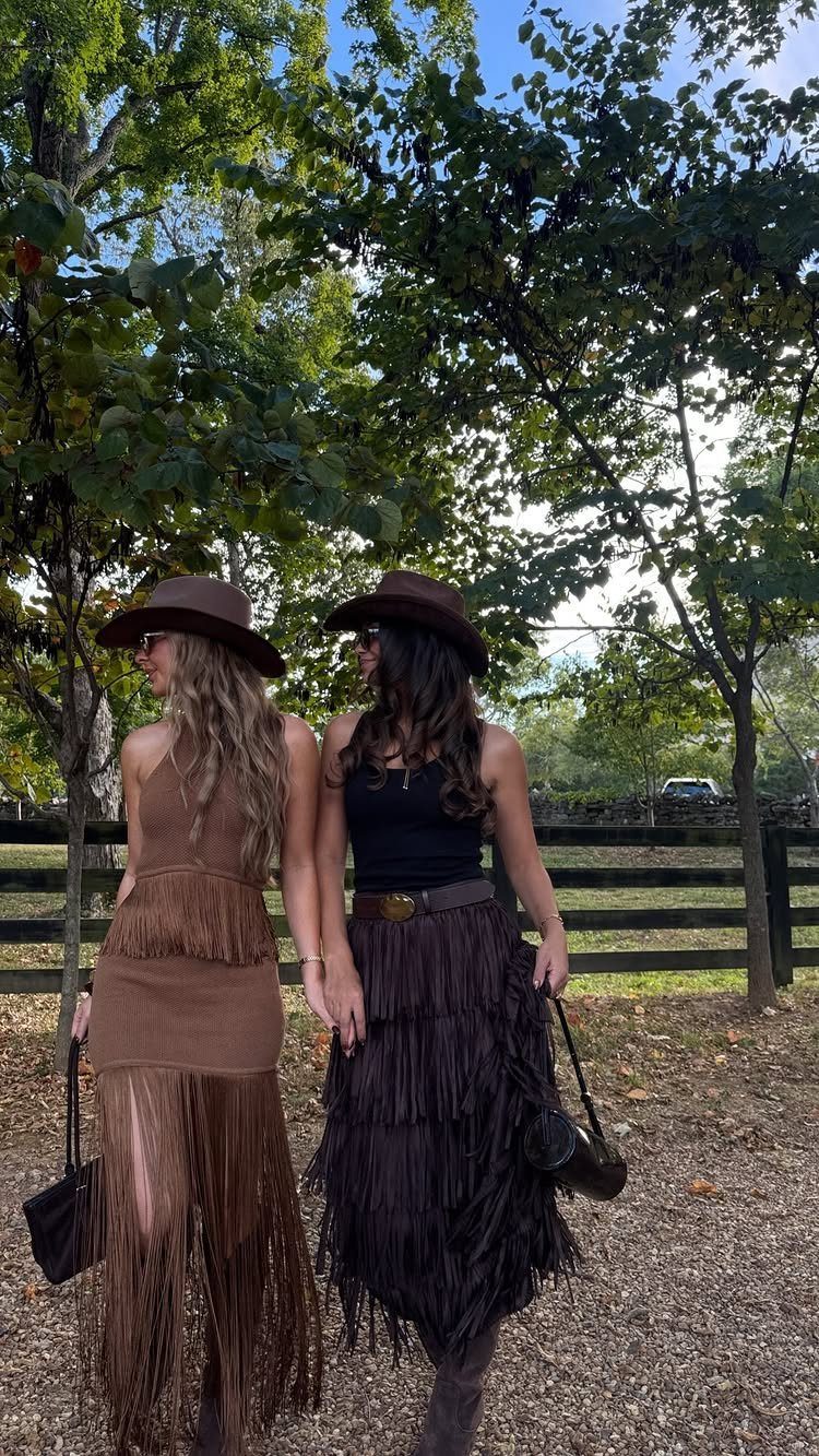 Two women in cowboy hats and fringe skirts walking through a wooded area.