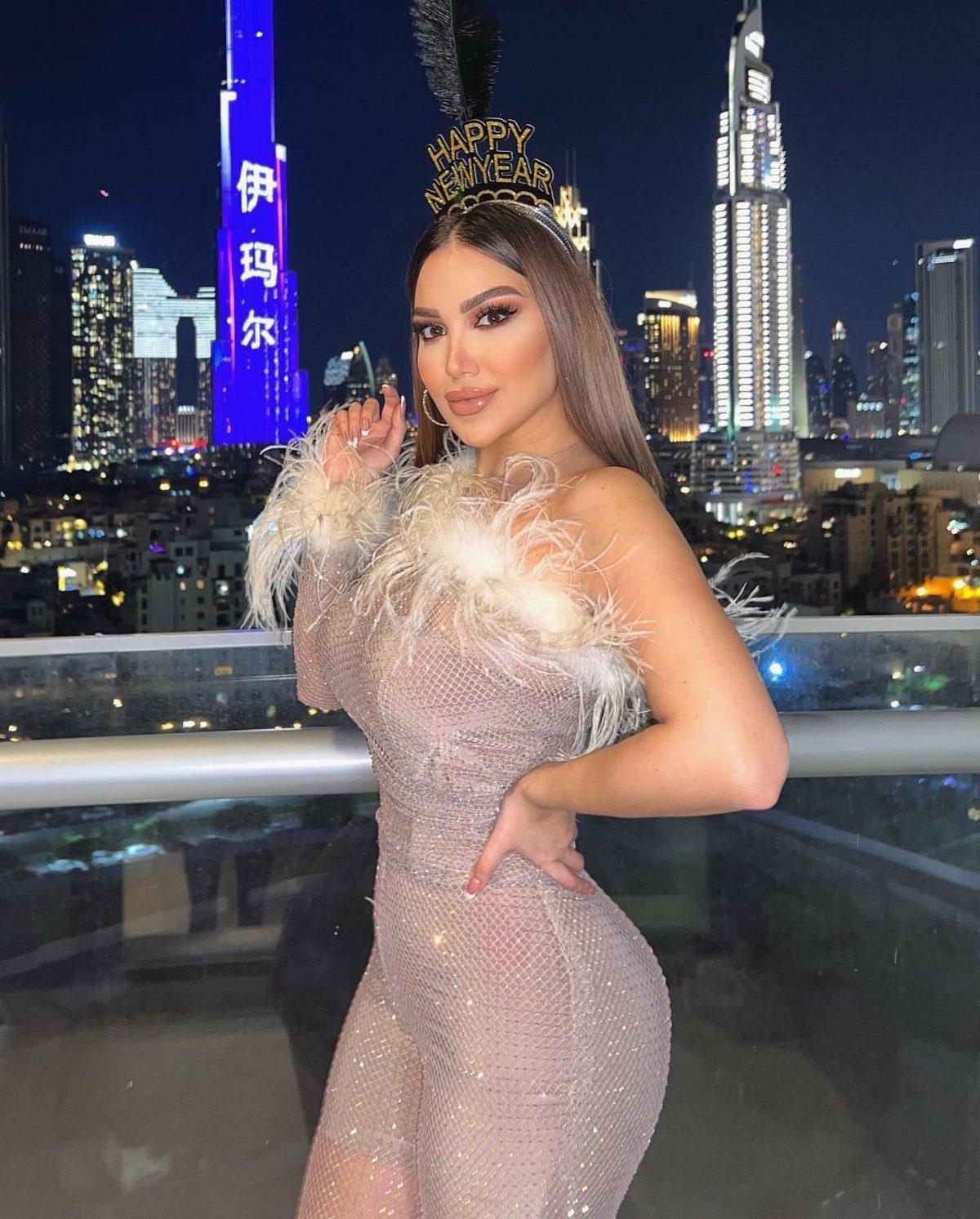 A woman in a sparkling feathered jumpsuit posing against a city skyline at night.