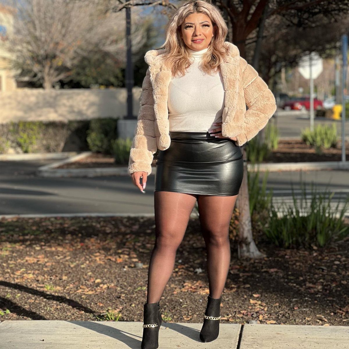 Woman wearing a faux fur jacket, white turtleneck, black leather skirt, and sheer black tights.