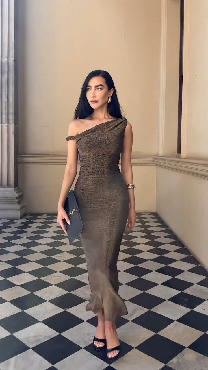 A woman posing in a long, one-shoulder metallic bronze dress on a checkered floor.