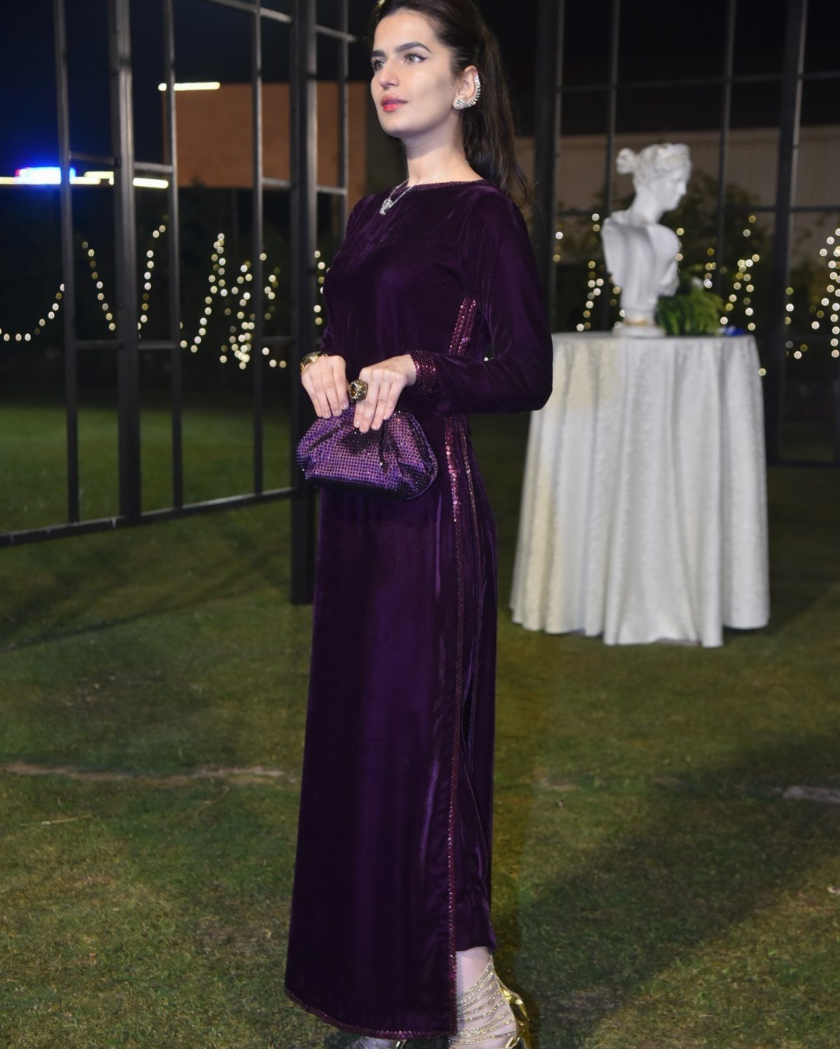 A woman posing in a long-sleeved, floor-length purple velvet dress and matching clutch.