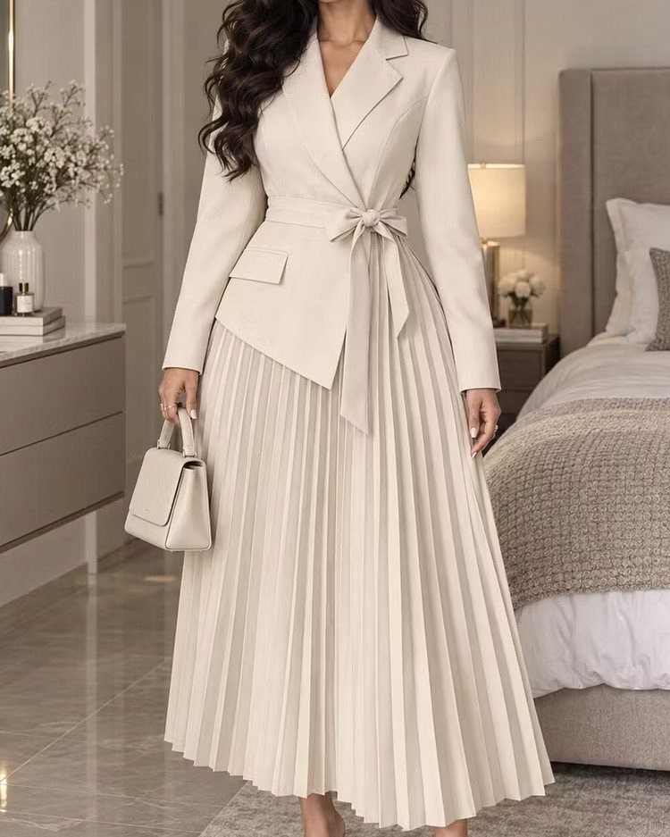 Woman in a cream blazer and long pleated skirt holding a small handbag.