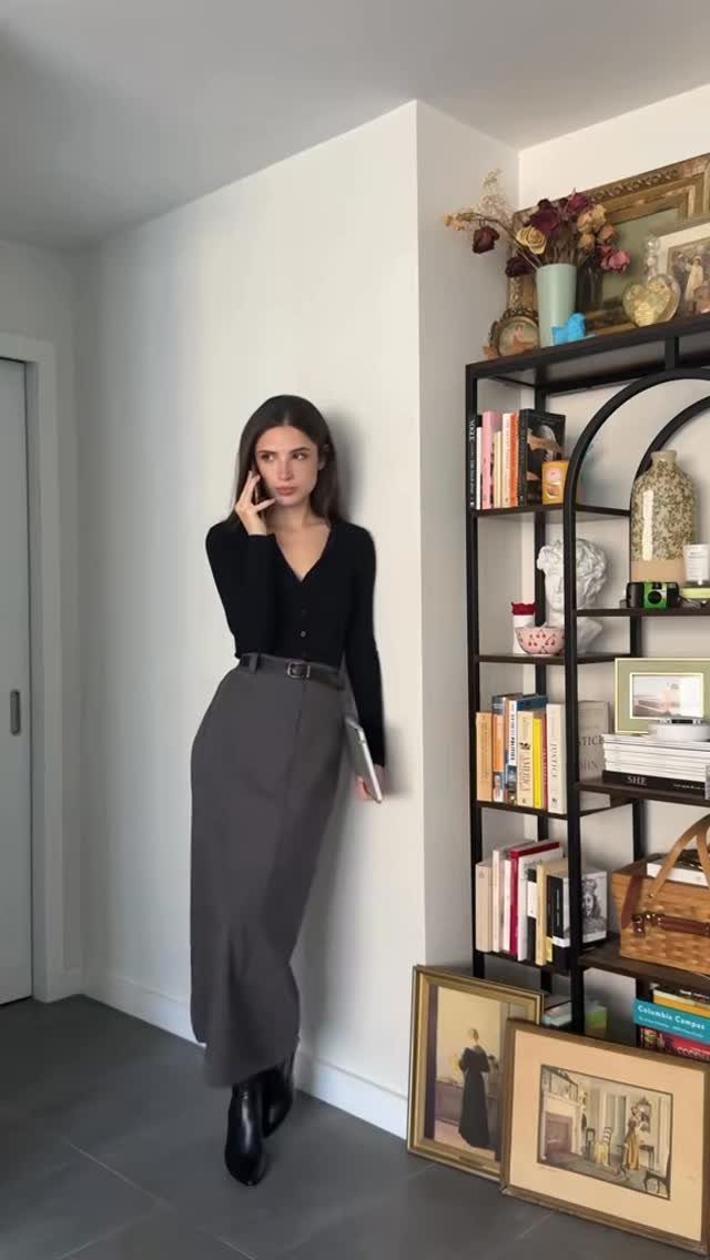 Woman wearing a black cardigan and long grey high-waisted skirt leaning against a white wall.