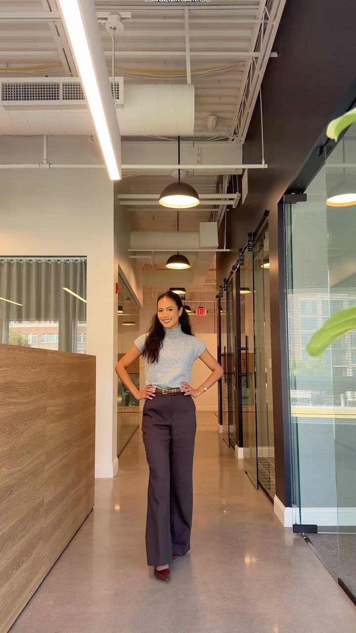 A woman posing in a modern office hallway wearing a grey knit top and dark trousers.