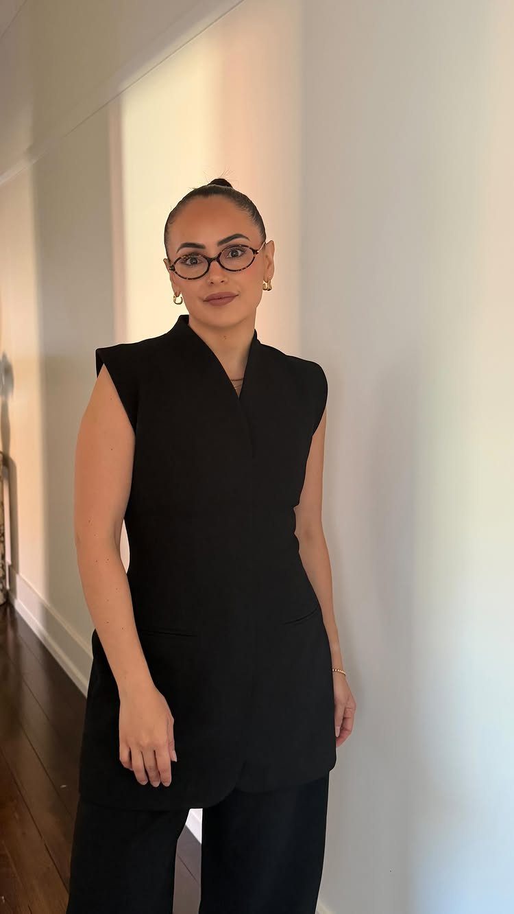 Woman in a black sleeveless tunic and trousers standing against a white wall.