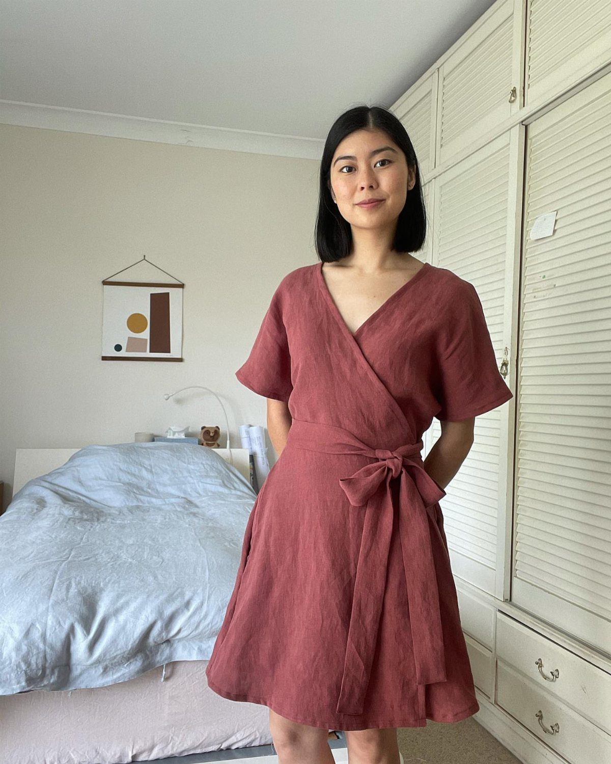 A woman posing in a mauve short-sleeved linen wrap dress in a bedroom setting.