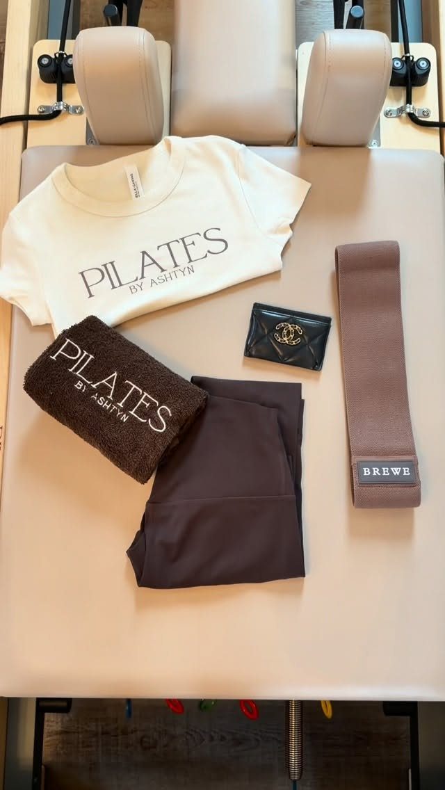 Cream t-shirt, dark leggings, and brown accessories laid out on a beige pilates reformer machine.