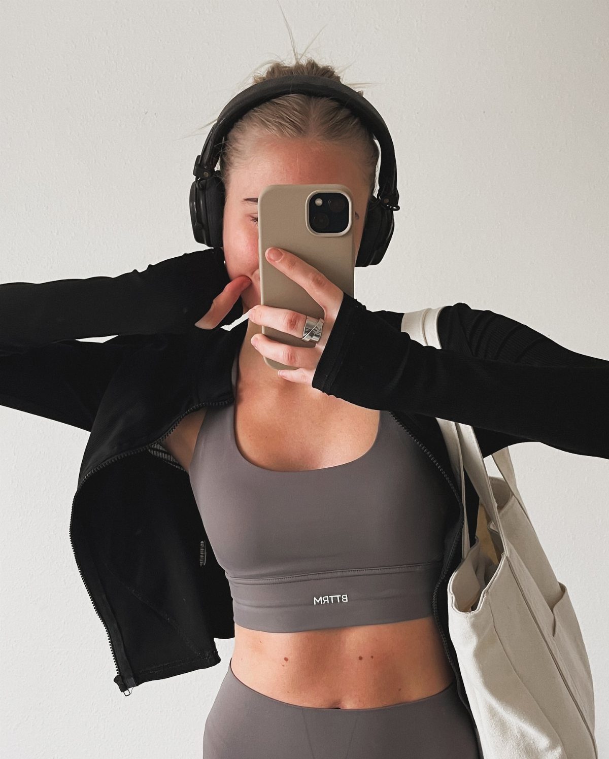 Woman in a grey sports bra and black jacket taking a mirror selfie.