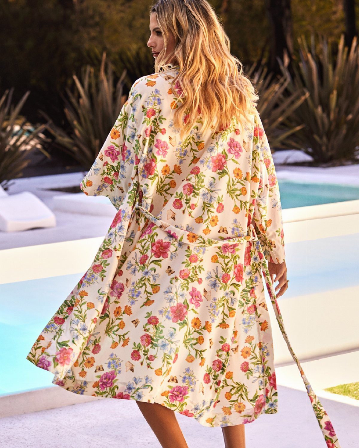A woman wearing a white floral print kimono jacket walking near a bright blue swimming pool.