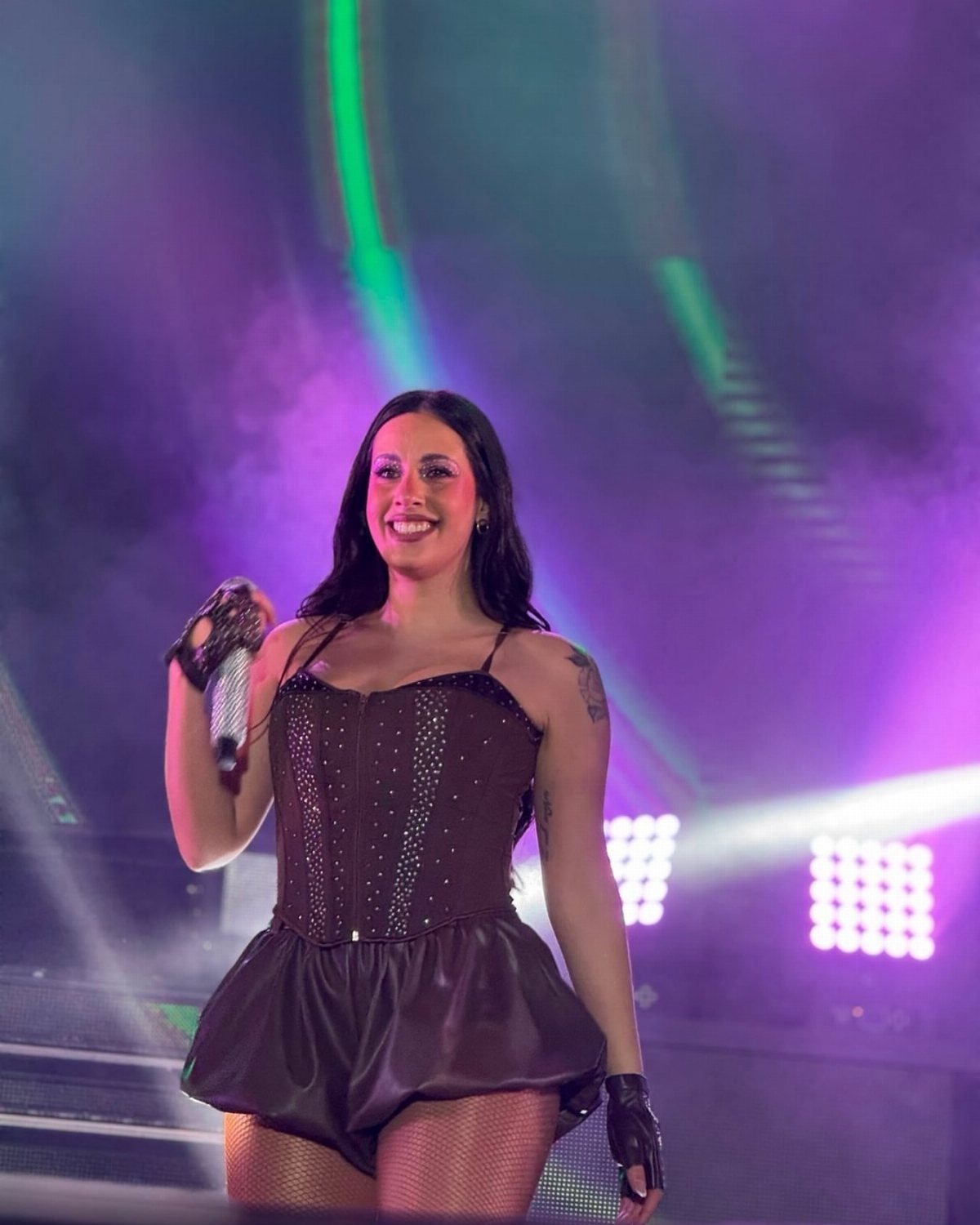 A woman performing on stage wearing a dark sequined corset and matching shorts under colorful lights.