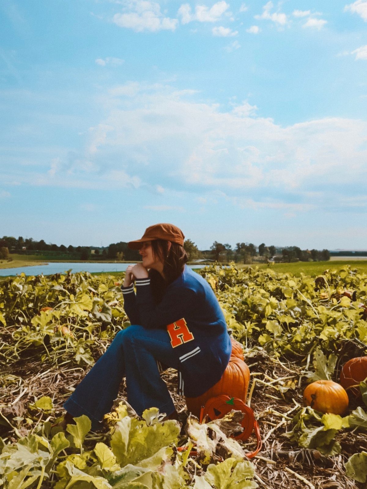 Woman in a blue cardigan and denim pants sitting in a pumpkin patch under a blue sky.