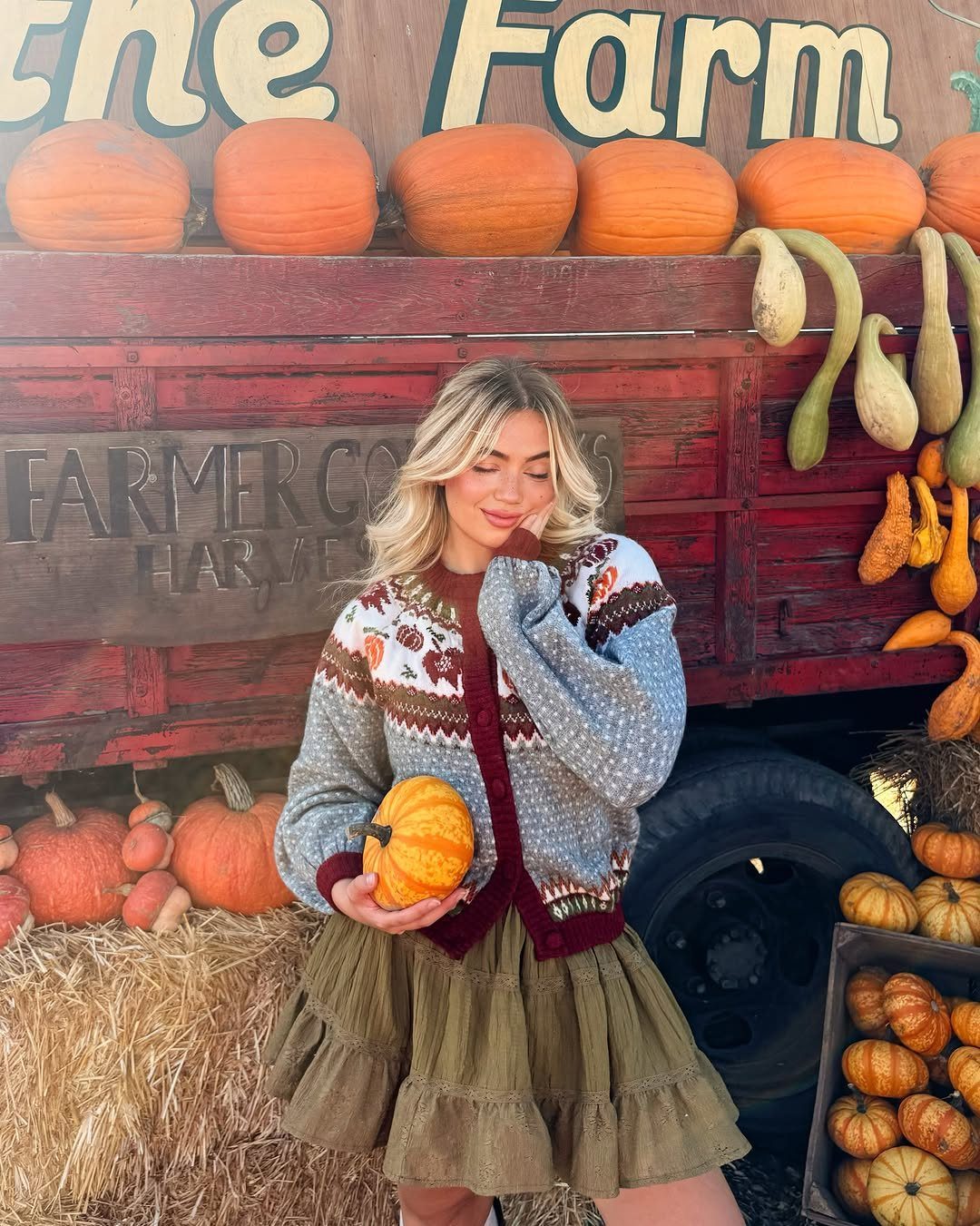 Blonde woman in a patterned cardigan and olive green ruffled skirt holding a pumpkin.