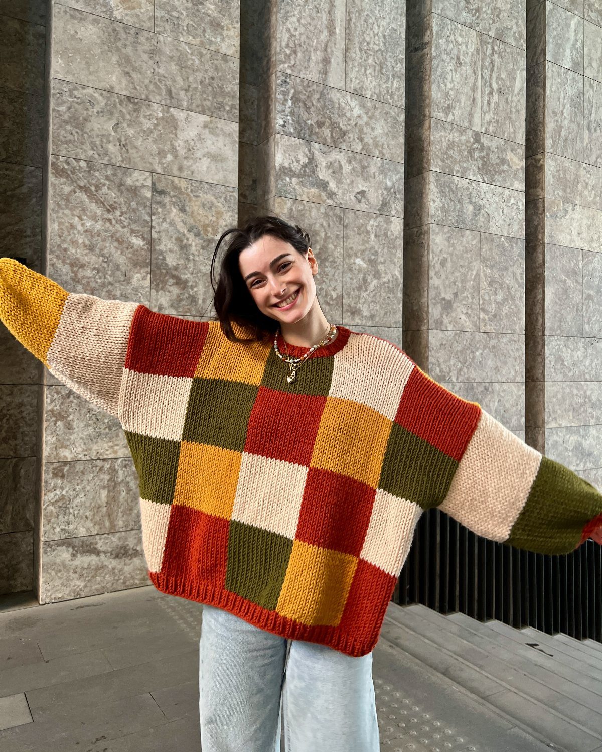 Smiling woman wearing a colorful checkered chunky knit sweater and light blue denim jeans.