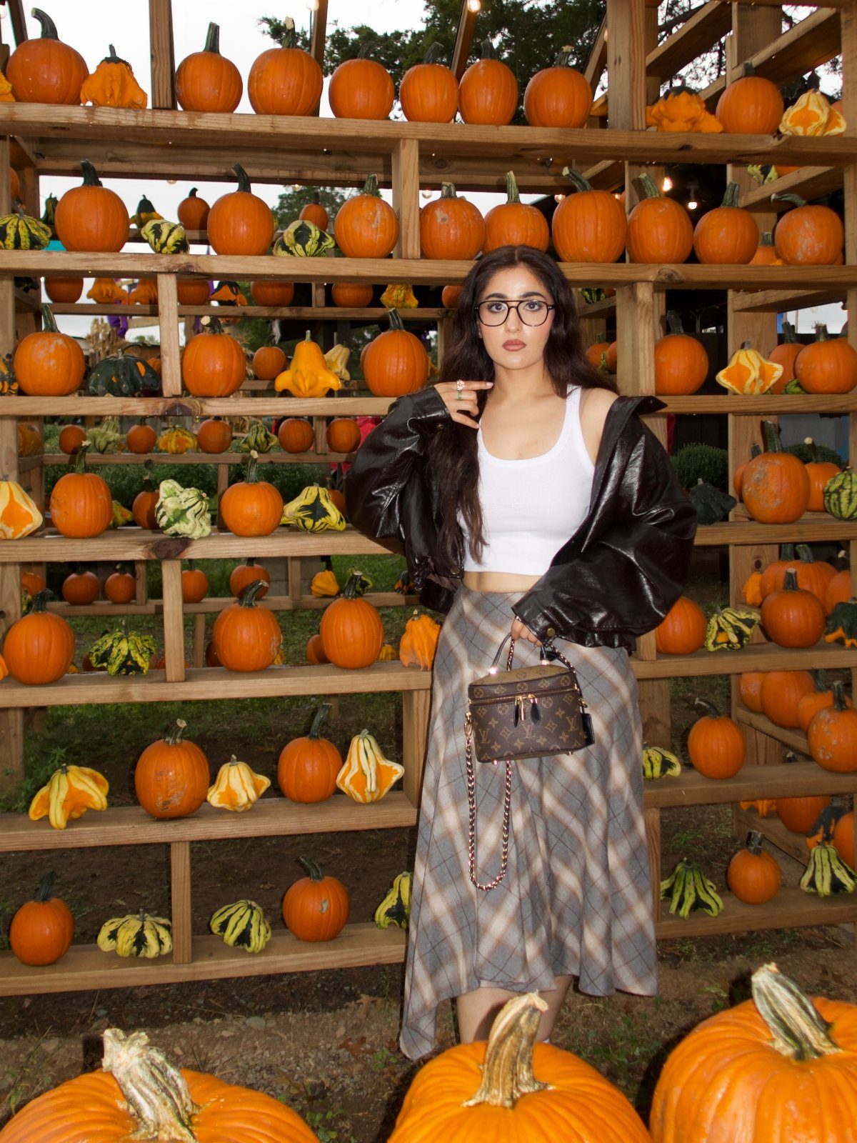 Woman in a black leather jacket and grey plaid skirt posing in front of pumpkin shelves.