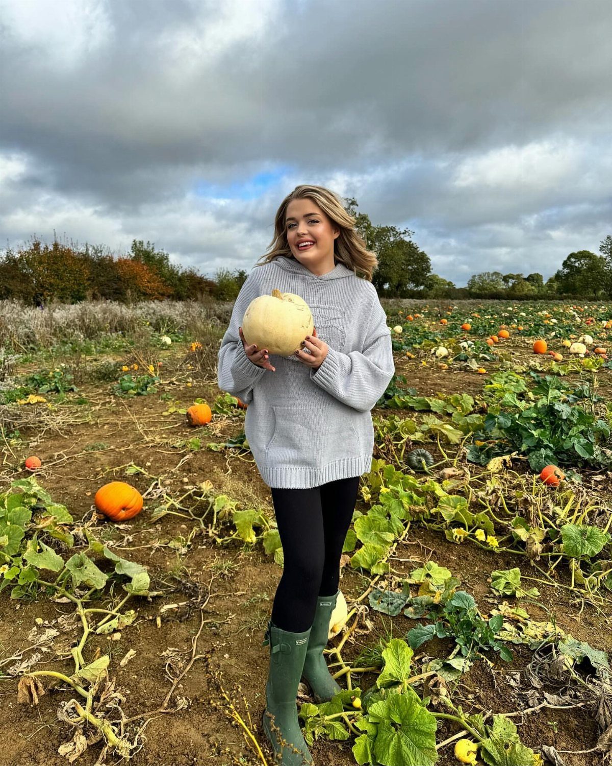 Smiling woman in a grey sweater and black leggings holding a white pumpkin in a field.