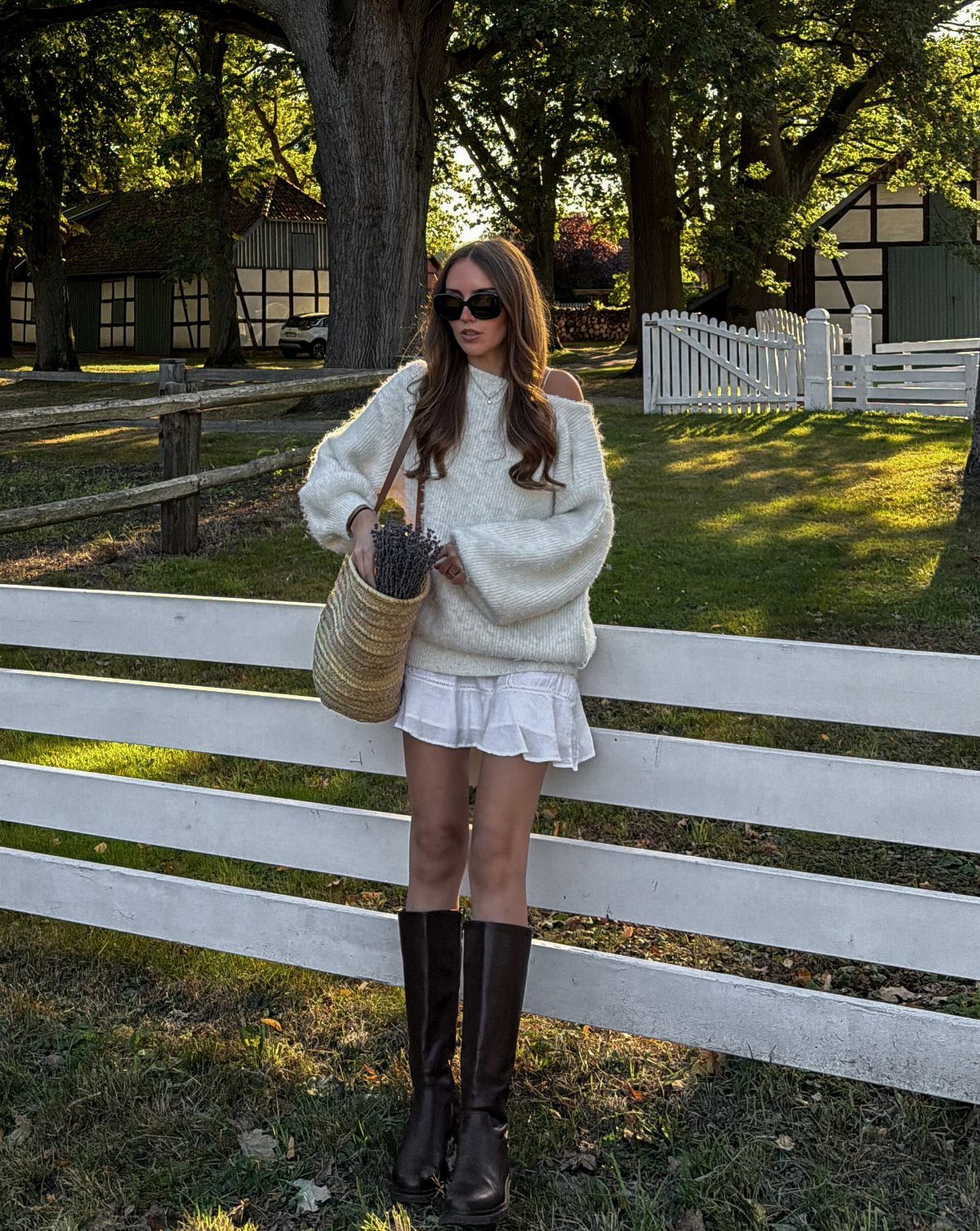 Woman wearing an oversized white sweater and ruffled skirt with tall brown boots standing by a fence.