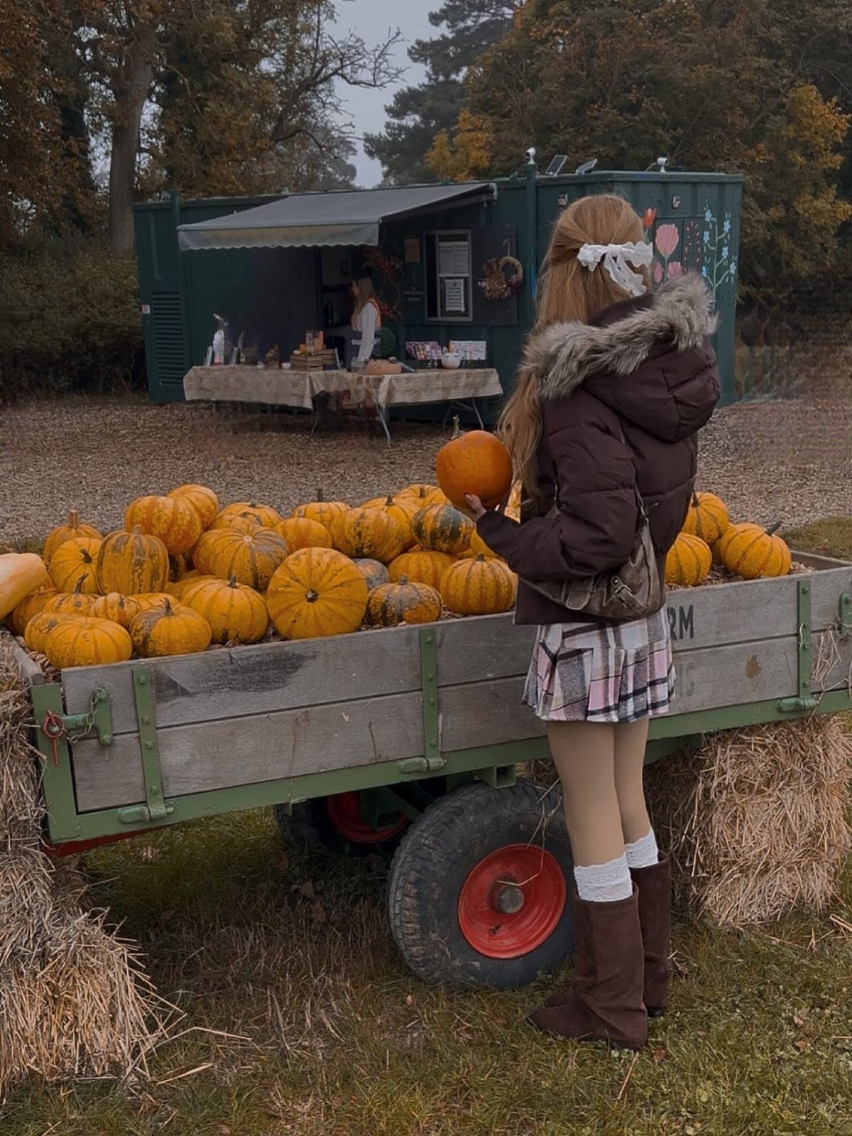 A person in a puffer jacket and plaid skirt holding a pumpkin near a wooden cart.
