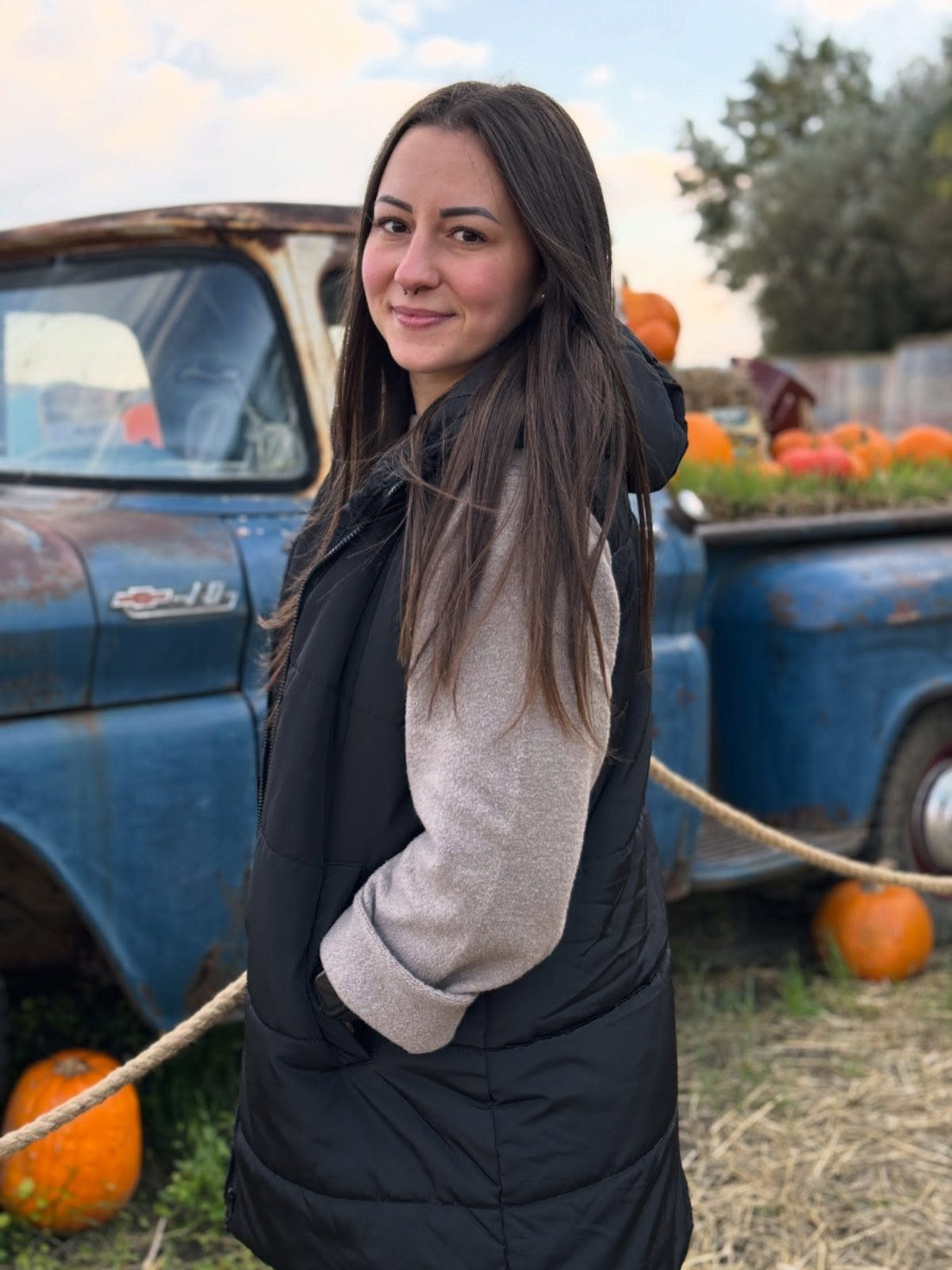 Woman wearing a black puffer vest and grey sweater standing near a blue truck and pumpkins.