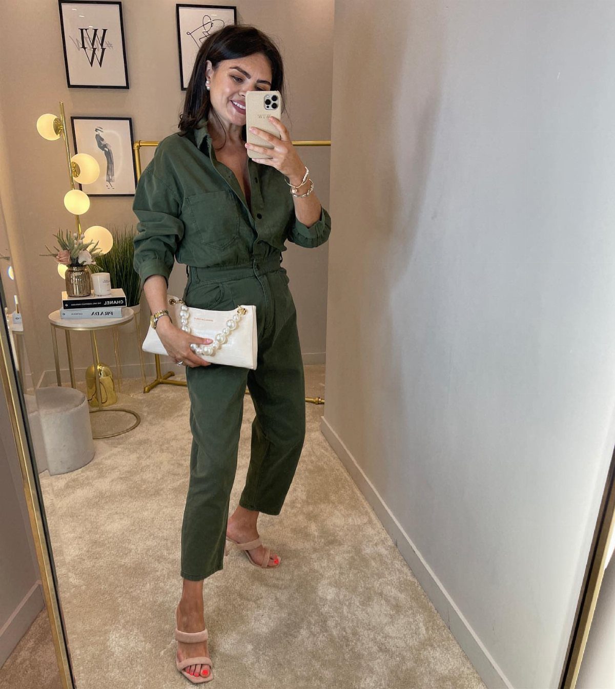 A woman posing in a mirror wearing an olive green jumpsuit and white handbag.