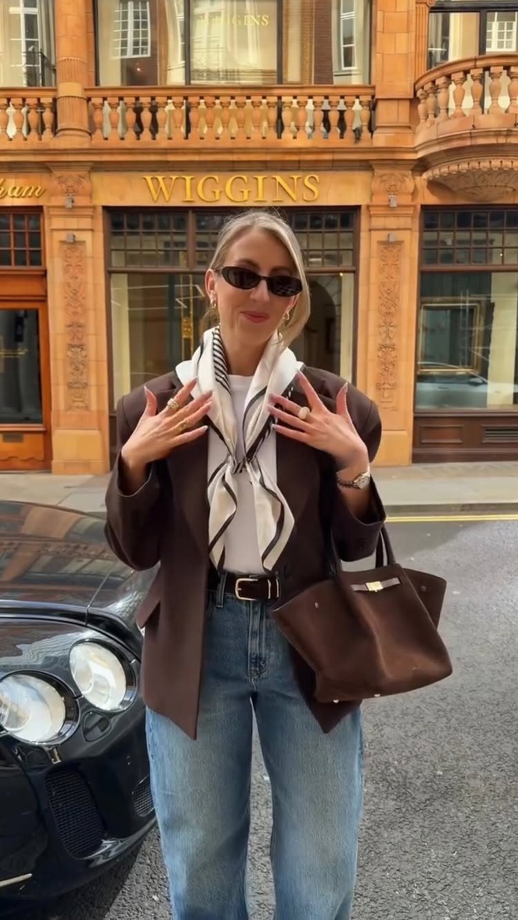 A woman wearing a brown blazer and a patterned lightweight scarf standing on a city street.