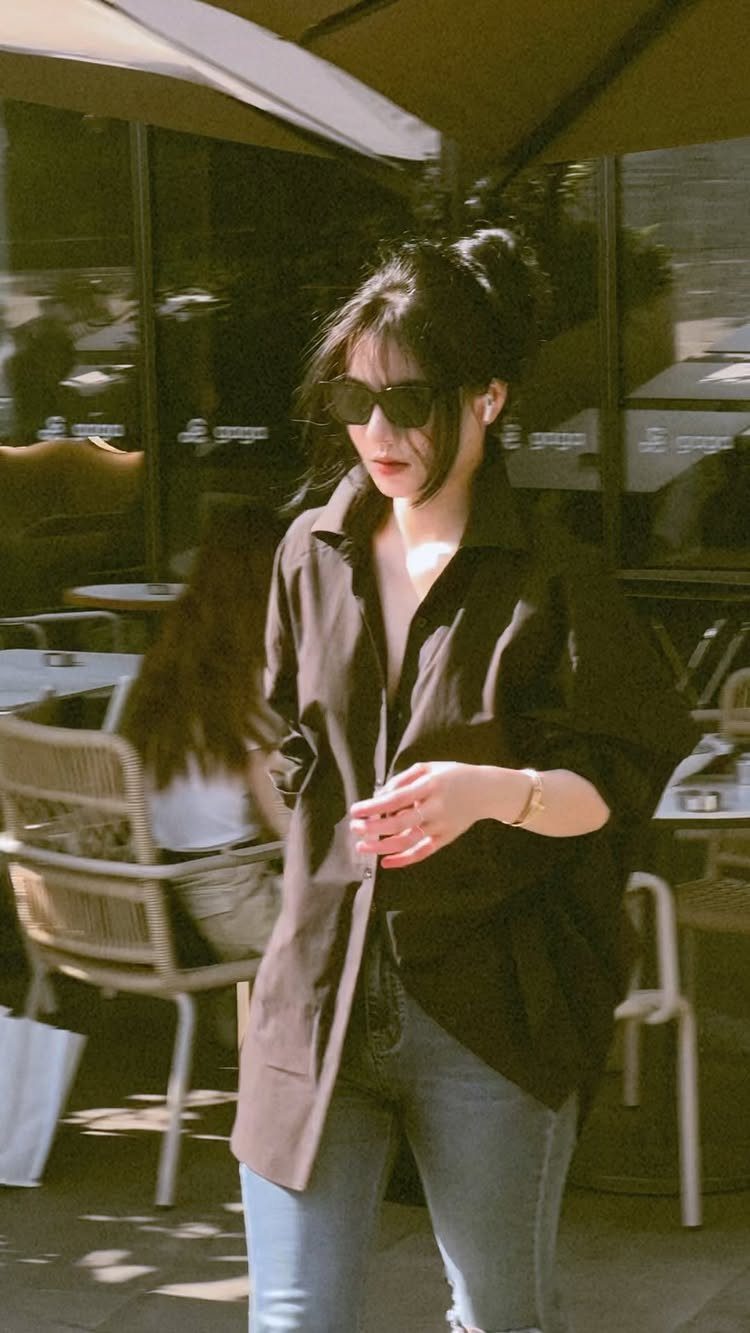A woman in an oversized dark shirt and blue jeans standing near an outdoor cafe.