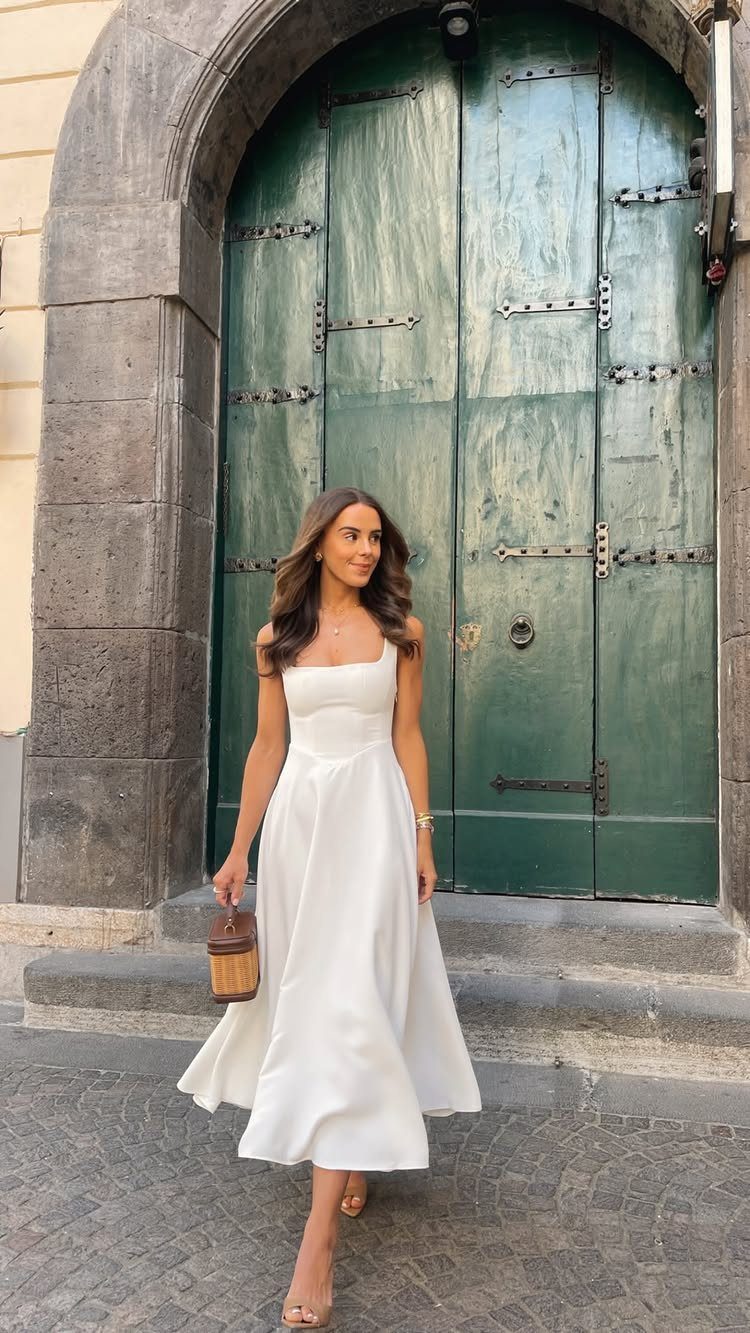 Woman in a white sleeveless midi dress walking in front of large green wooden doors.