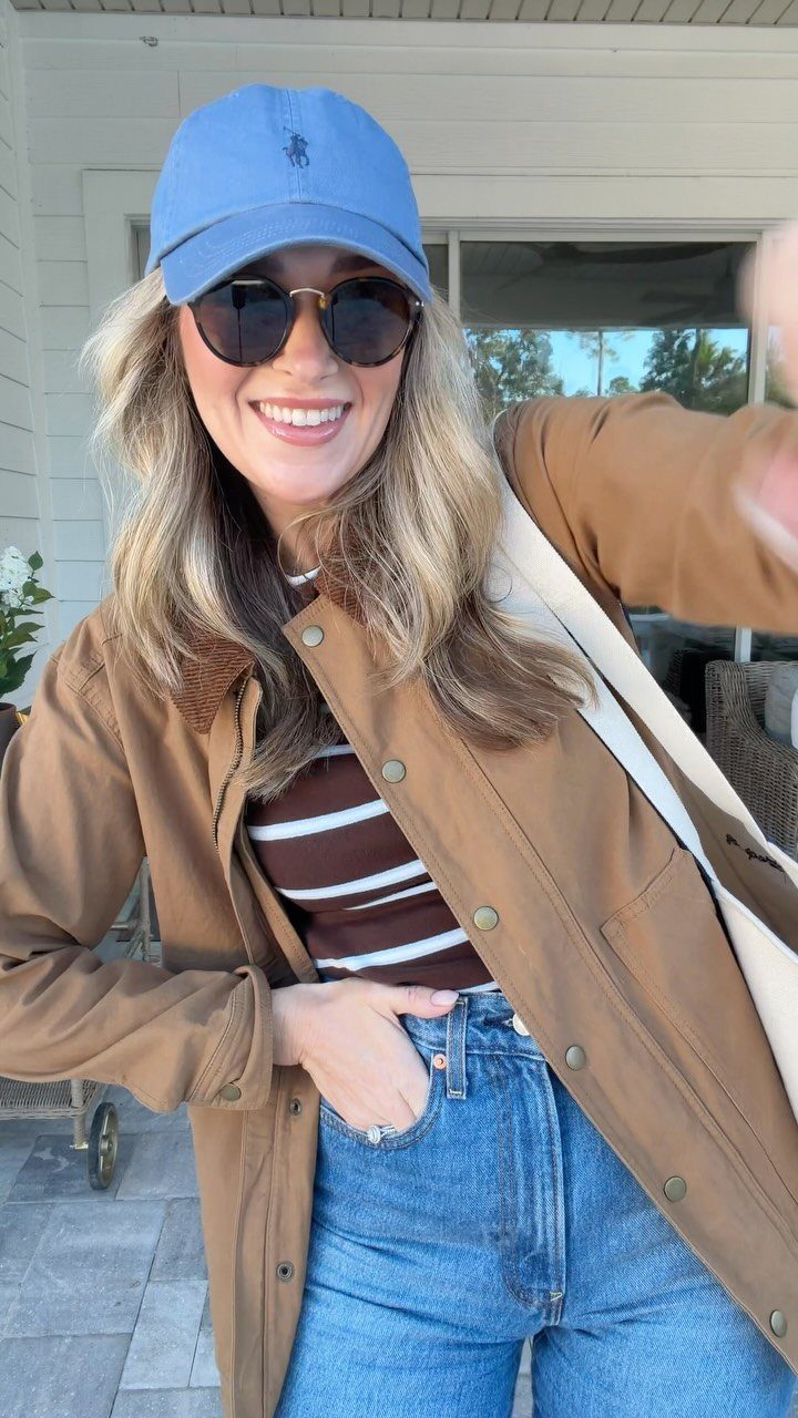 Smiling woman wearing a blue baseball cap, sunglasses, brown jacket, and blue jeans outdoors.