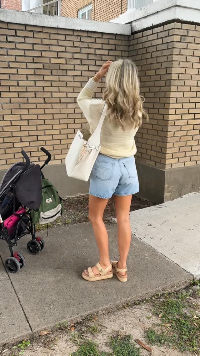 Blonde woman wearing a cream top and denim shorts standing next to a stroller.