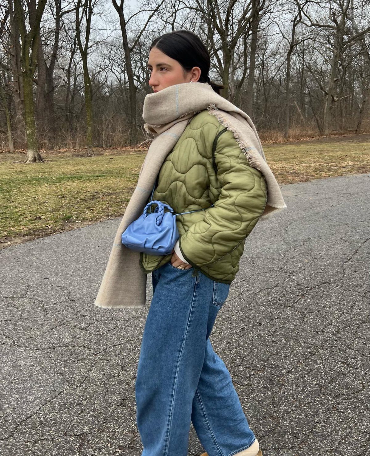 Woman wearing a green quilted jacket, blue jeans, and a large beige scarf walking outdoors.