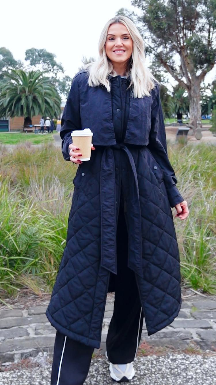 Blonde woman in a long black quilted coat and athletic pants holding a coffee cup.