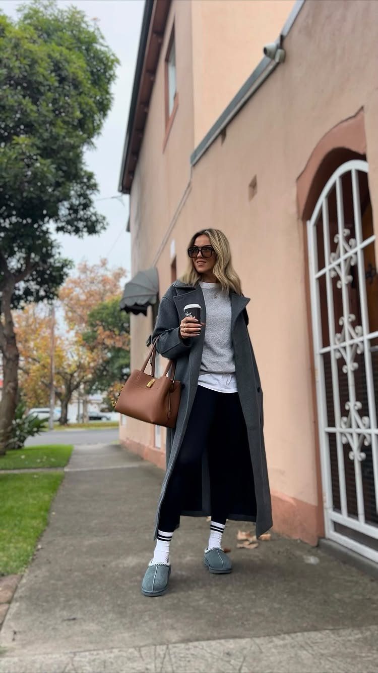 Woman in a long grey coat, black leggings, and grey clogs walking on a sidewalk.