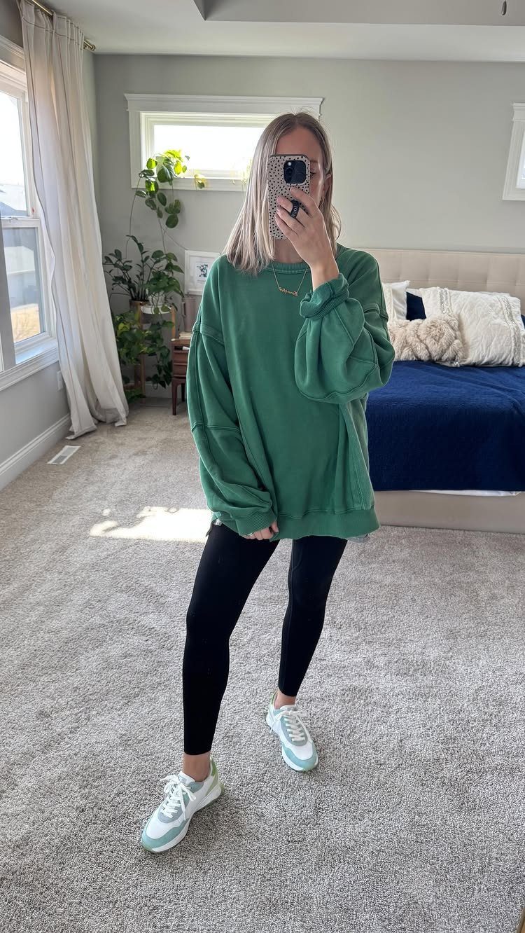 Woman in a green sweatshirt, black leggings, and white sneakers posing in a bedroom.