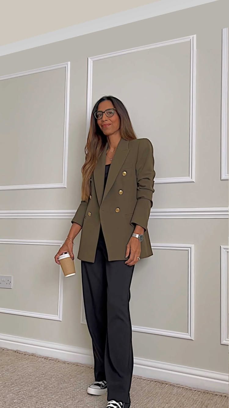 Woman posing in an olive green blazer and dark tailored trousers holding a coffee cup.