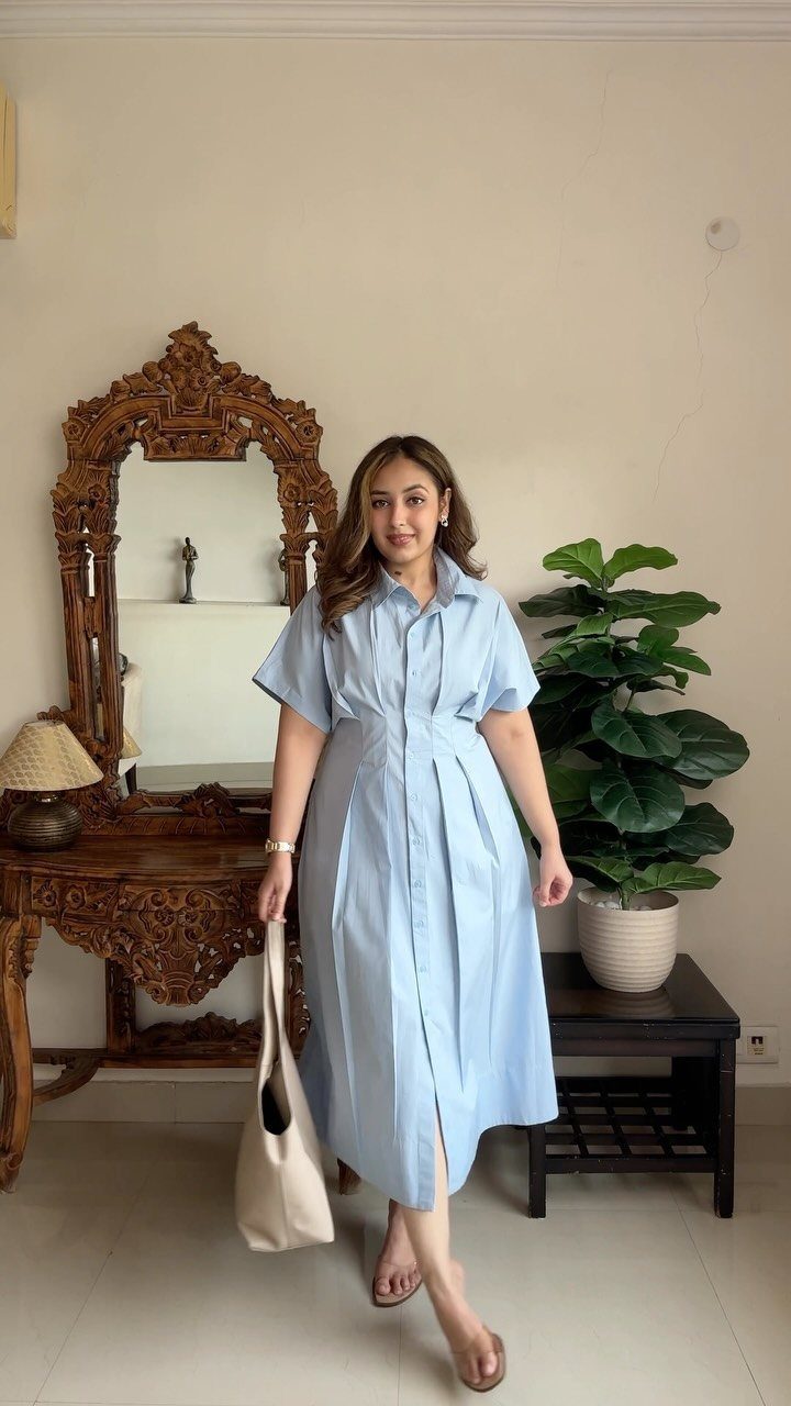 Woman in a light blue midi shirtdress standing in a room with an ornate mirror.