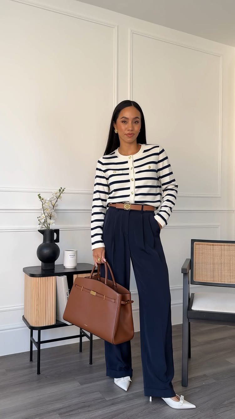 Woman posing in a striped top, navy trousers, and white heels while holding a brown leather handbag.