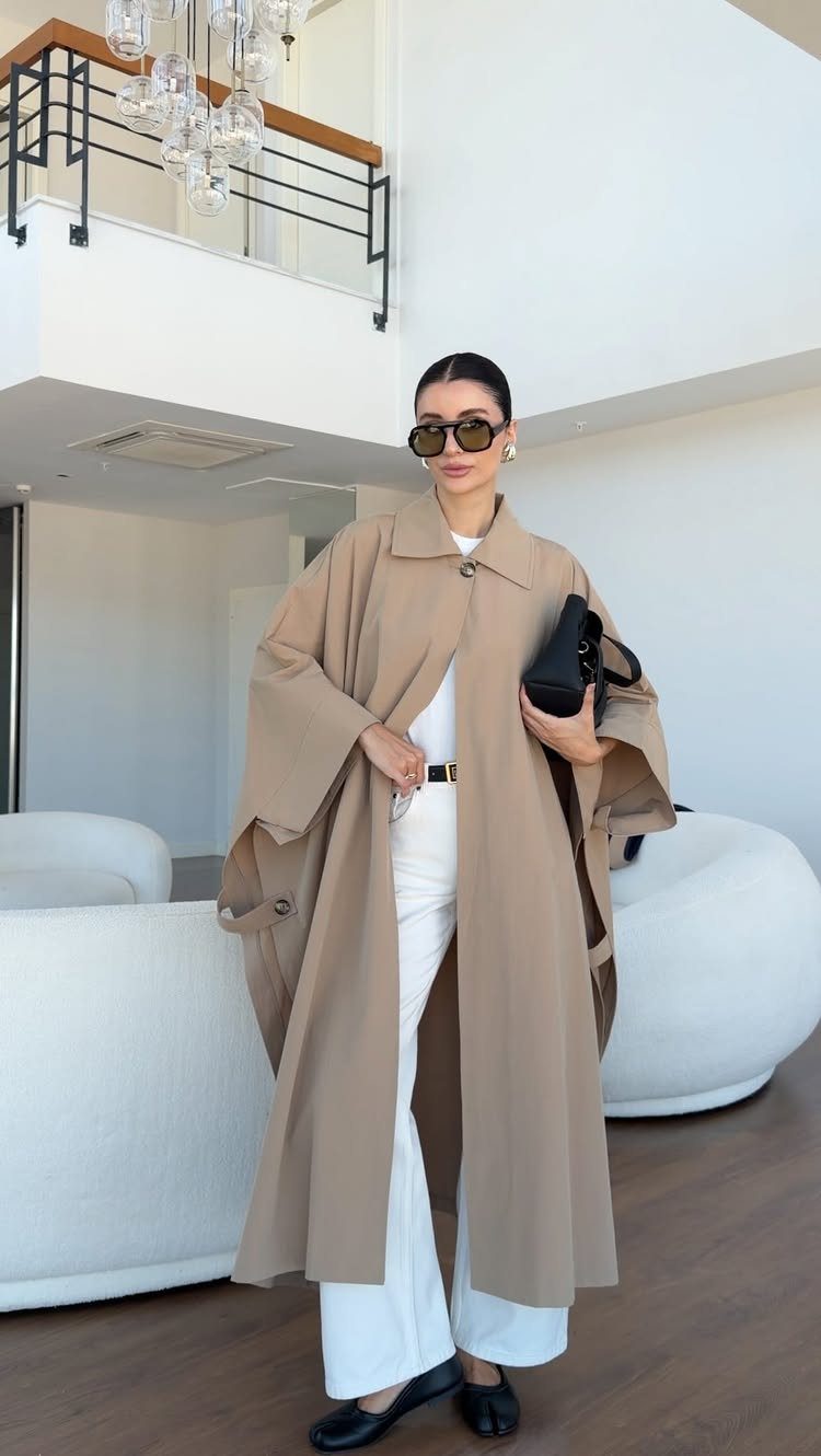 Woman in a tan trench coat, white trousers, and sunglasses standing in a modern room.