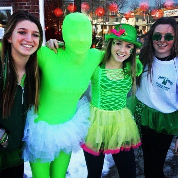 Four young women smiling and posing in various green St. Patrick's Day themed outfits outdoors.