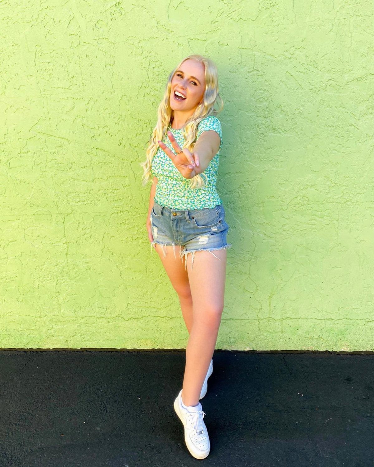 Smiling blonde woman wearing a green patterned shirt and denim shorts against a lime green wall.