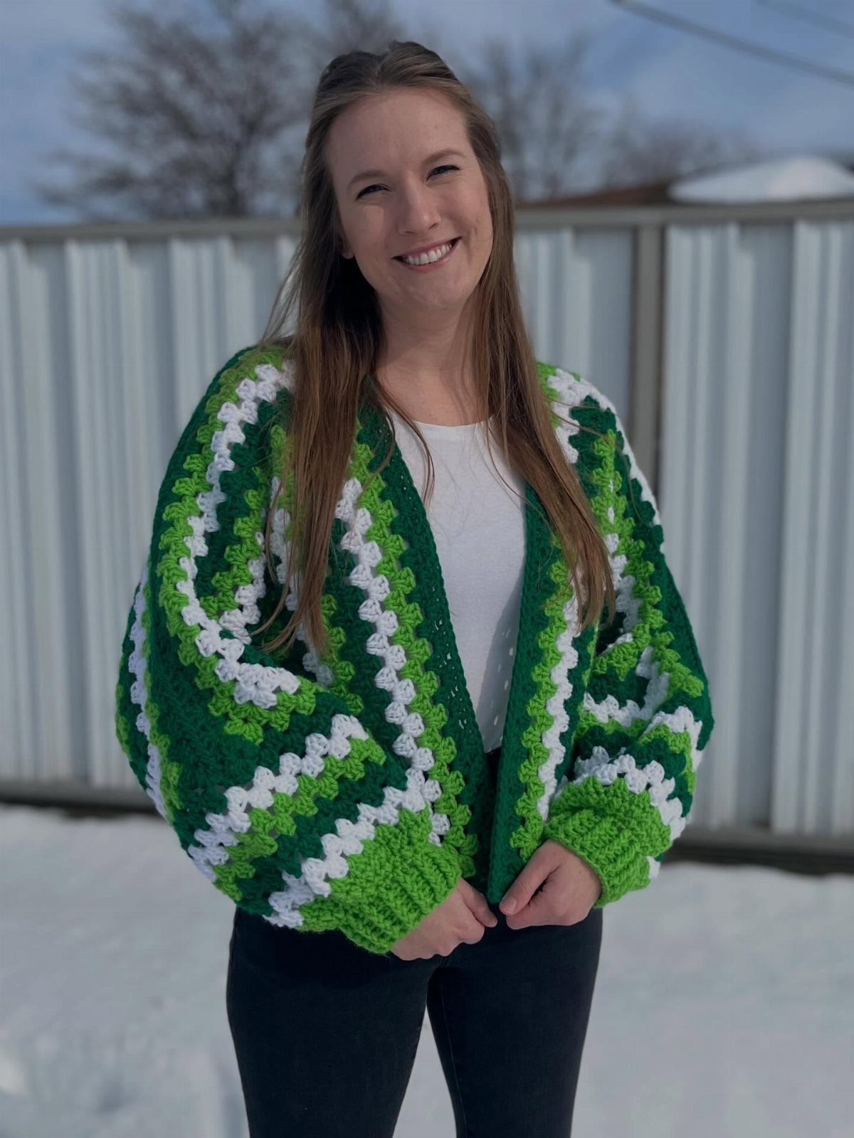 A smiling woman wearing a green and white striped crochet cardigan and dark pants in the snow.