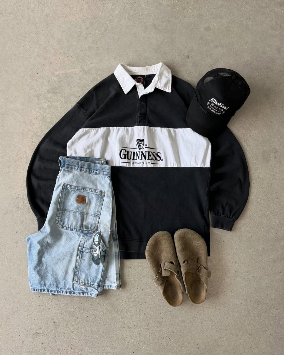 Guinness polo shirt, denim shorts, black cap, and tan sandals laid out on concrete.