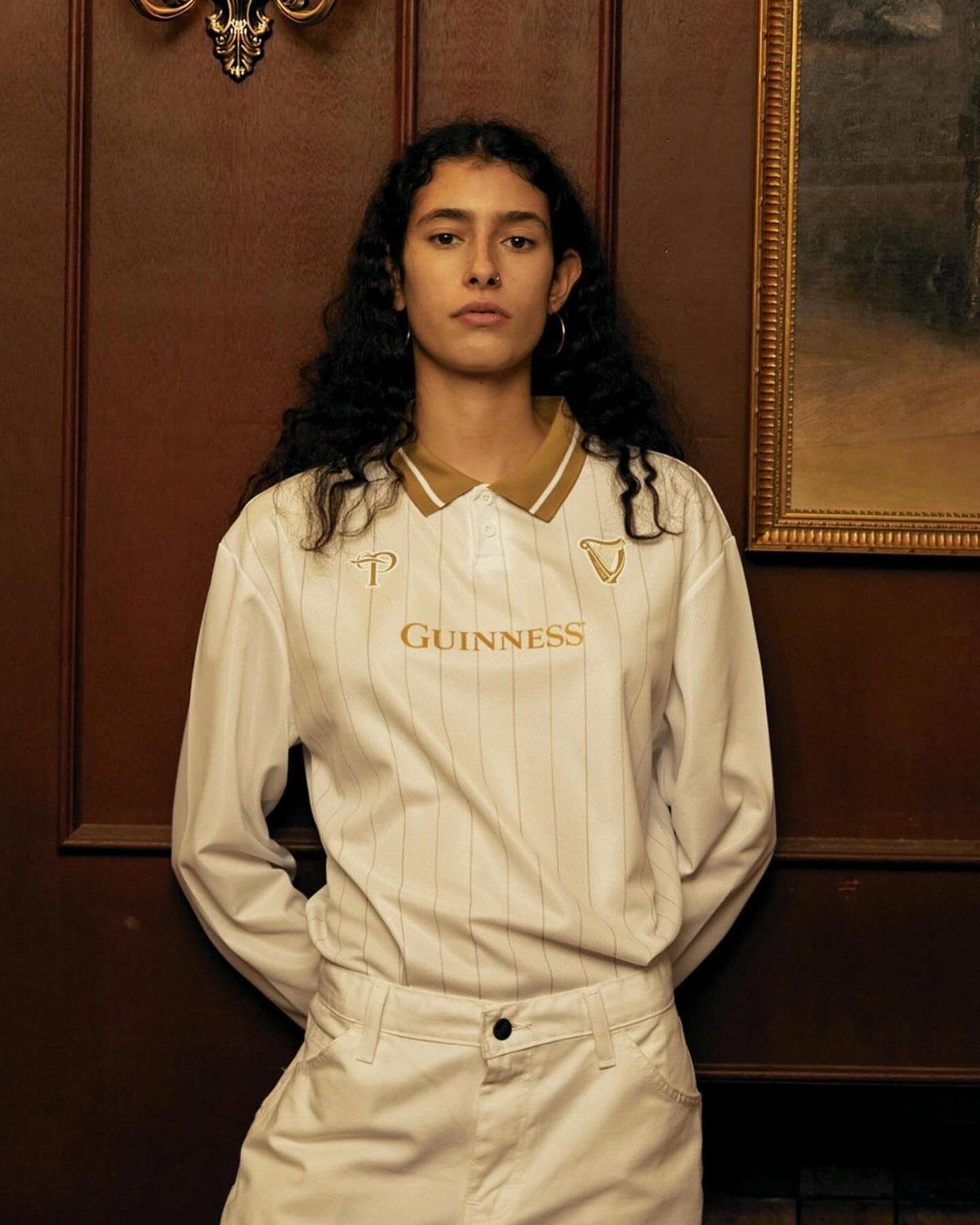 Woman wearing a white Guinness rugby shirt and white pants standing against a dark wood wall.
