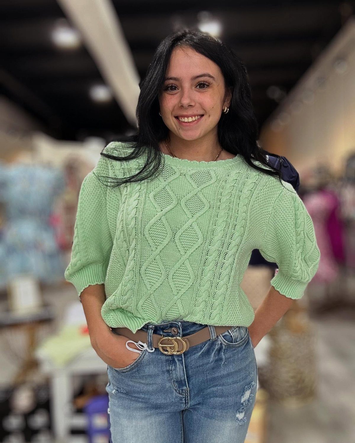 A smiling young woman wearing a light green cable knit sweater and blue denim jeans.