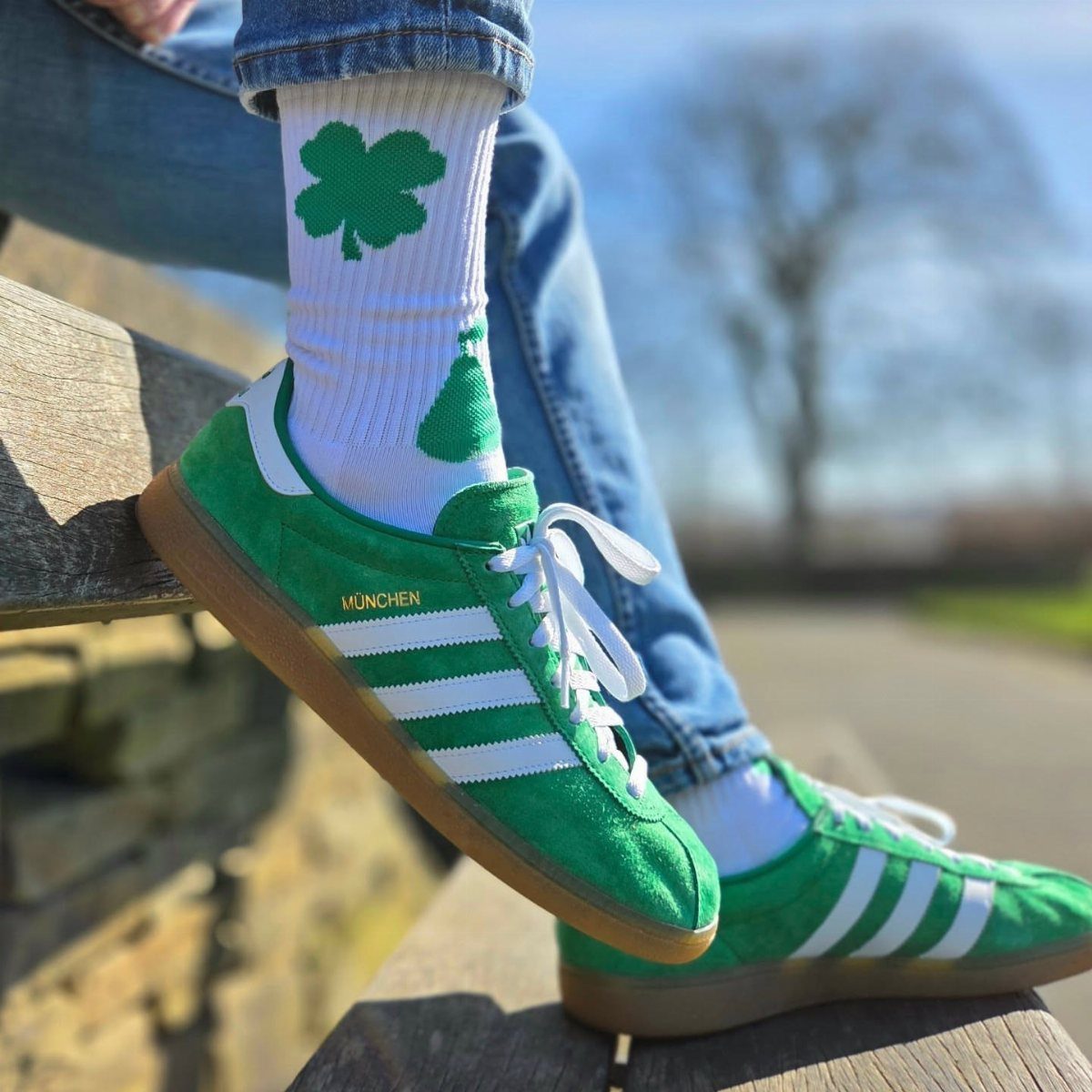 Green suede sneakers with white stripes and shamrock patterned socks worn with blue denim jeans.