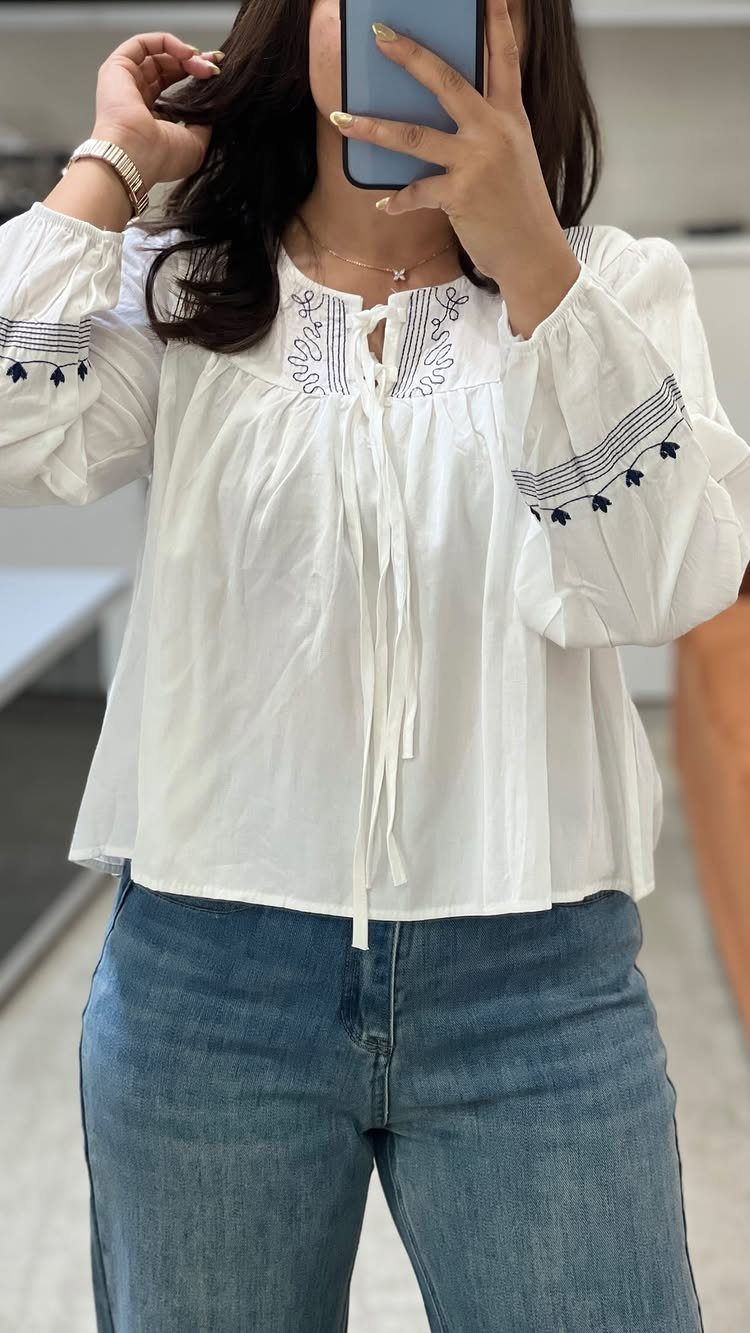 Woman in a white embroidered blouse and blue jeans taking a mirror selfie.